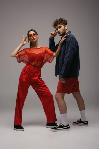 Full length of stylish woman in red outfit and sunglasses posing near man on grey — Stock Photo