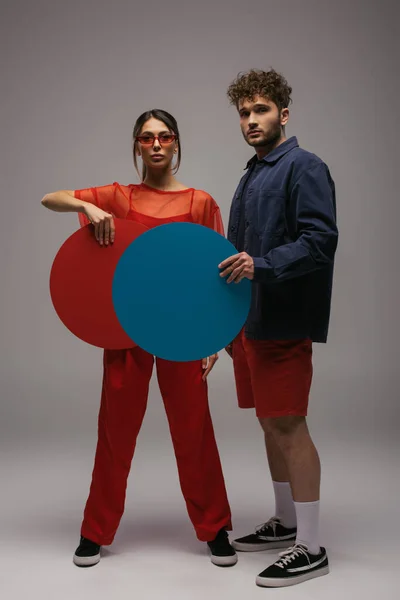 Full length of stylish couple in trendy outfits and sunglasses holding round shape carton on grey — Photo de stock