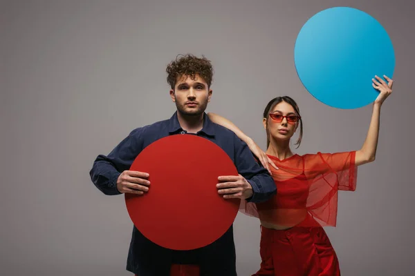 Stylish models in trendy blue and red outfits holding round shape carton isolated on grey - foto de stock