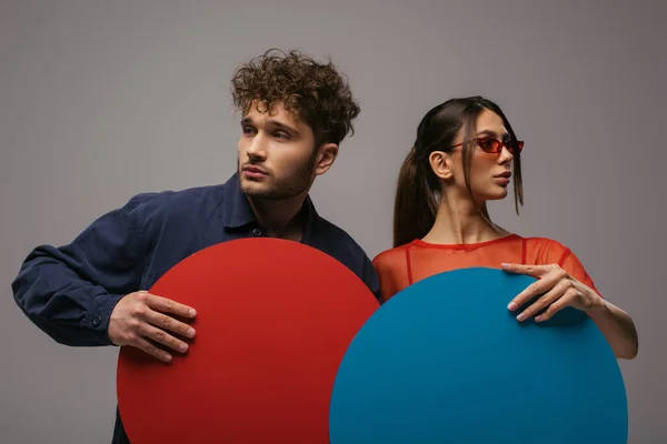 Young couple in trendy blue and red outfits holding round shape carton and looking away isolated on grey — стоковое фото