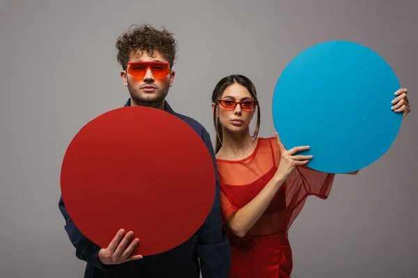 Young couple in blue and red outfits and sunglasses holding round shape carton isolated on grey - foto de stock
