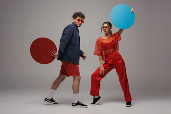 Full length of young couple in trendy outfits and sunglasses holding round shape carton on grey — Photo de stock