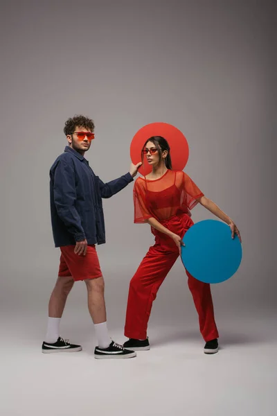 Full length of stylish couple in blue and red outfits holding round shape carton on grey - foto de stock
