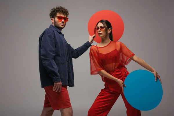 Jovem casal elegante em roupas azuis e vermelhas segurando forma redonda caixa isolada no cinza — Fotografia de Stock