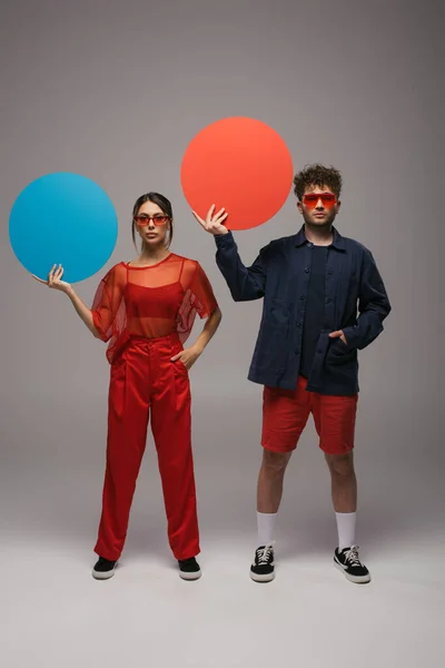 Full length of couple in trendy outfits holding round shape carton and posing with hands in pockets on grey - foto de stock