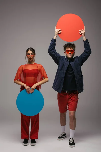 Full length of young stylish couple in blue and red outfits posing with round shape carton on grey — Photo de stock