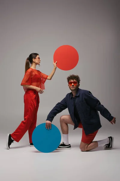 Full length of young couple in blue and red outfits posing with round shape carton on grey - foto de stock