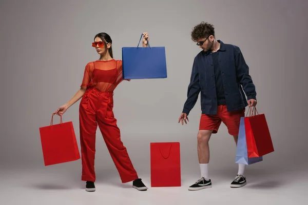 Full length of young couple in trendy outfits holding shopping bags on grey - foto de stock