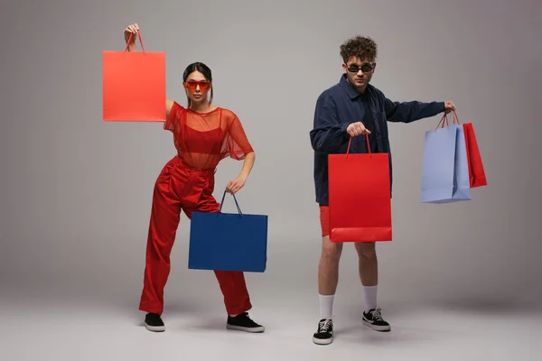 Full length of stylish young couple in trendy outfits holding shopping bags on grey — Stock Photo