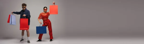 Comprimento total de jovem casal em roupas da moda segurando sacos de compras em cinza, banner — Fotografia de Stock