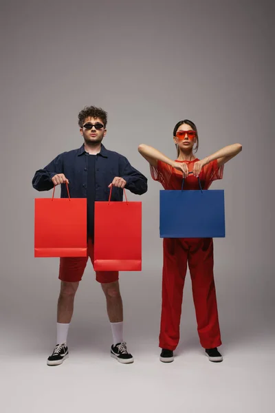 Full length of stylish couple in trendy outfits and sunglasses holding shopping bags on grey — стоковое фото