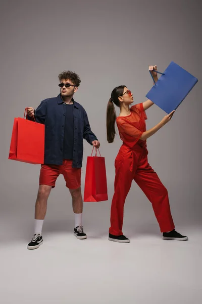 Full length of young models in trendy outfits and sunglasses holding shopping bags on grey - foto de stock