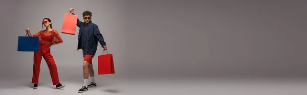 Full length of young couple in trendy outfits and sunglasses holding shopping bags while posing on grey, banner — Photo de stock