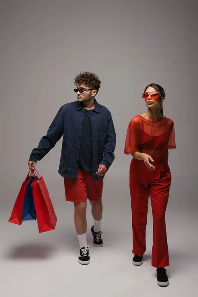 Full length of young couple in trendy outfits and sunglasses walking with shopping bags on grey — Stockfoto