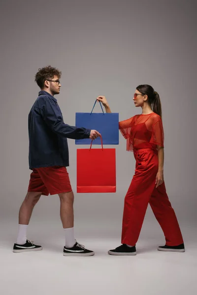 Side view of young couple in trendy outfits and sunglasses holding shopping bags on grey — Foto stock