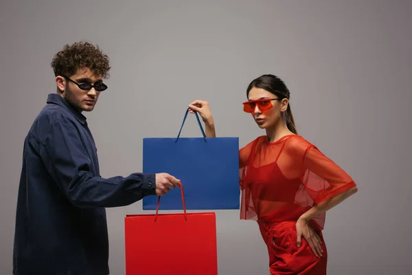 Alla moda giovane coppia in abiti alla moda e occhiali da sole che tengono borse della spesa isolate sul grigio — Foto stock