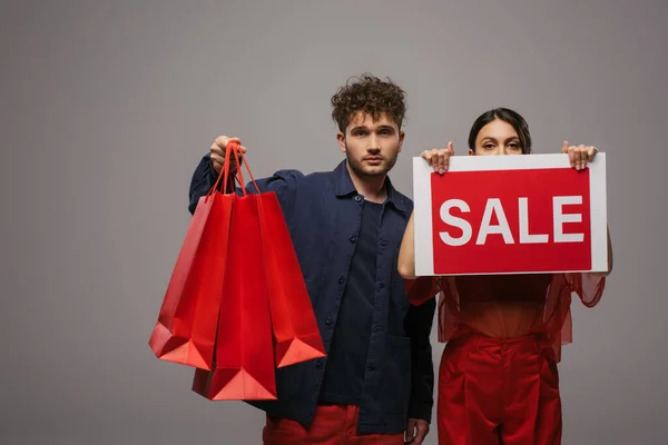 Young couple in trendy outfits holding shopping bags and paper with sale lettering isolated on grey — Fotografia de Stock