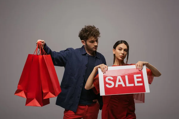 Paar in trendigen Outfits mit Einkaufstaschen und Papier mit Verkaufsbuchstaben isoliert auf grau — Stockfoto
