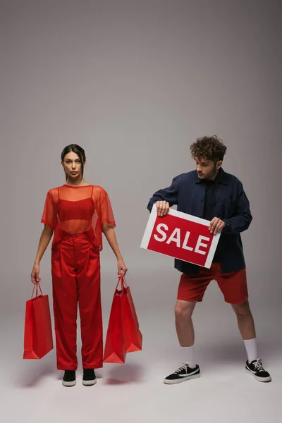 Full length of young models in trendy outfits holding shopping bags and paper with sale lettering on grey — Photo de stock