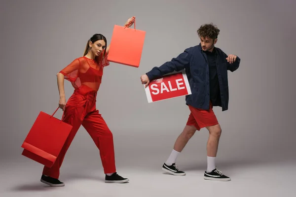 Models in trendigen Outfits mit Einkaufstaschen und Papier mit Verkaufsbuchstaben auf grau — Stockfoto