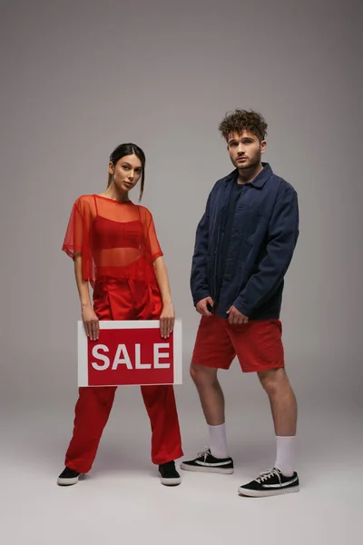 Full length of young woman holding paper with sale lettering near man on grey — стоковое фото