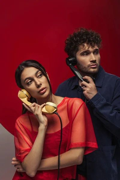 Young woman and curly man holding retro handsets while posing on red and grey — стоковое фото