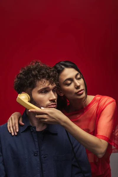 Junge Frau hält Retro-Hörer in der Nähe von Ohr des lockigen Mannes auf rot — Stockfoto