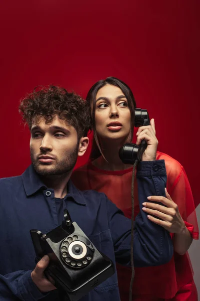 Curly man holding handset and retro telephone near young woman on red - foto de stock