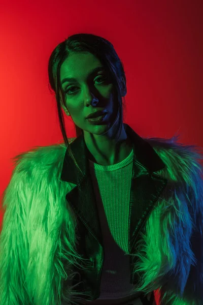 Portrait of stylish young woman in faux fur jacket looking at camera on red — Stockfoto