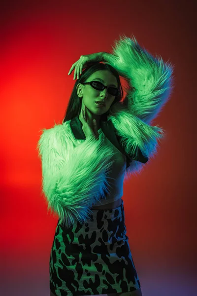 Stylish young woman in faux fur jacket and sunglasses posing on red — Photo de stock