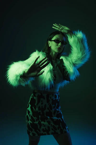 Стильная молодая женщина в искусственном меховом пиджаке и солнцезащитных очках жестикулирует, позируя на синем — стоковое фото