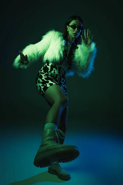 Full length of stylish young woman in faux fur jacket and sunglasses posing on blue — Photo de stock