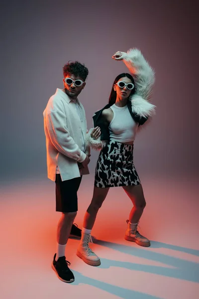 Full length of trendy woman in white faux fur jacket and skirt with animal print posing near man in sunglasses on grey — Photo de stock