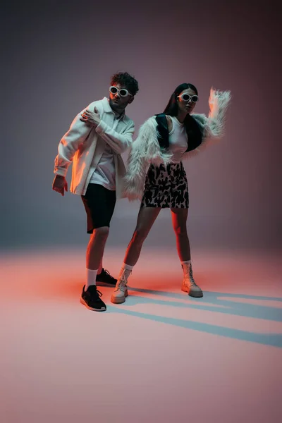 Full length of fashionable woman in faux fur jacket and skirt with animal print posing near man in sunglasses on grey — Photo de stock