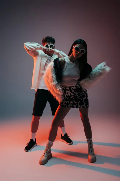 Full length of fashionable woman in white faux fur jacket and skirt with animal print posing with man in sunglasses on grey — Stock Photo