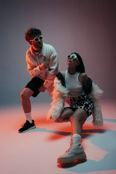 Full length of stylish woman in white faux fur jacket and skirt with animal print posing with man in sunglasses on grey — Photo de stock