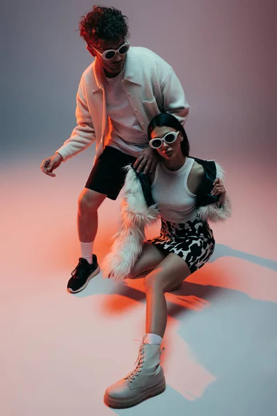 Full length of stylish woman in faux fur jacket and skirt with animal print posing with curly man in sunglasses on grey — Photo de stock