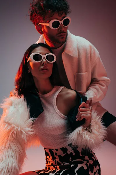 Uomo riccio e donna alla moda in occhiali da sole e giacca di pelliccia sintetica in posa sul grigio — Foto stock