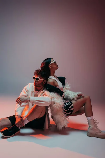 Stylish man in sunglasses and woman in faux fur jacket sitting while posing on pink and grey — Photo de stock