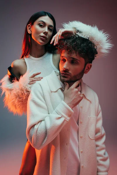 Young woman in faux fur jacket gesturing near thoughtful man posing on grey — Foto stock