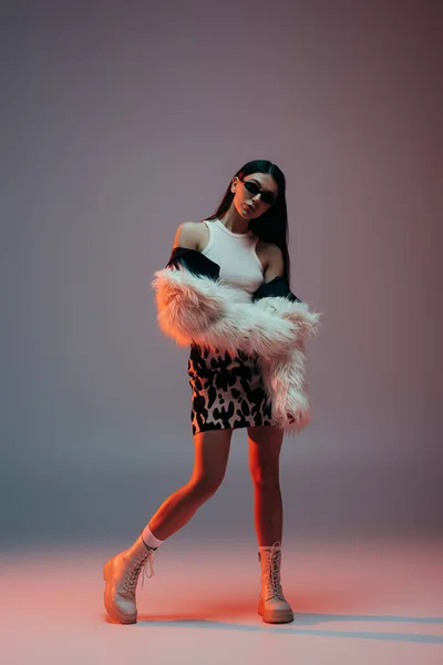 Full length of fashionable young woman in skirt with animal print and white faux fur jacket posing on grey - foto de stock