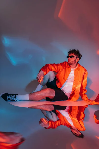 Volle Länge des lockigen und stilvollen Mannes mit Sonnenbrille posiert, während er im Studio mit buntem Licht sitzt — Stockfoto