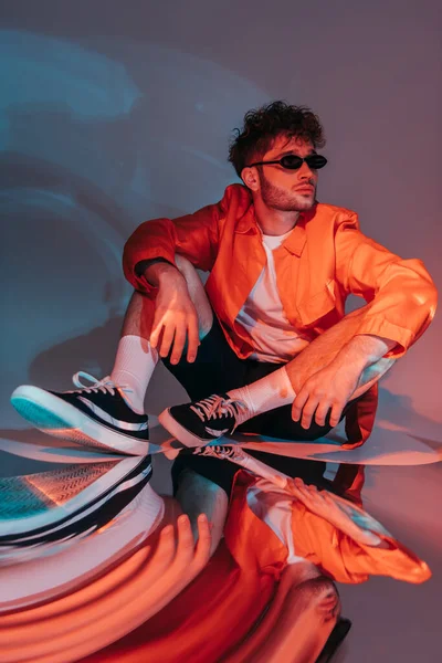 Full length of trendy man in sunglasses posing while sitting in studio with colorful light - foto de stock