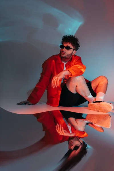 Full length of curly man in sunglasses posing while sitting on grey with colorful light — Stock Photo