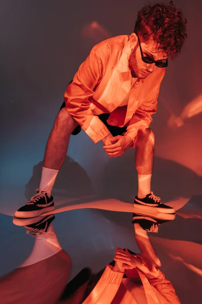 Full length of curly man in sunglasses looking at reflection on grey with colorful light — Stockfoto