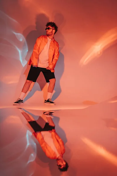 Full length of curly and trendy man in sunglasses posing while looking away on orange with colorful light — Photo de stock