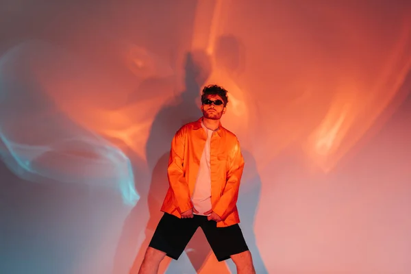 Curly and trendy man in sunglasses posing while looking and camera on red and blue with lighting — Stock Photo