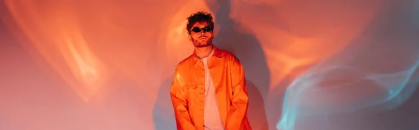 Curly and trendy man in sunglasses posing while looking and camera on red and blue with lighting, banner — Photo de stock