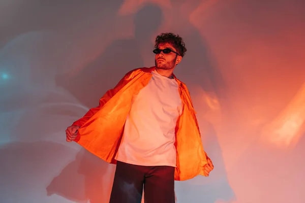 Curly and trendy man in sunglasses and orange shirt posing in studio with lighting — стоковое фото