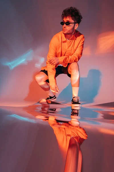 Full length of curly young man in sunglasses posing while sitting on grey with colorful light - foto de stock